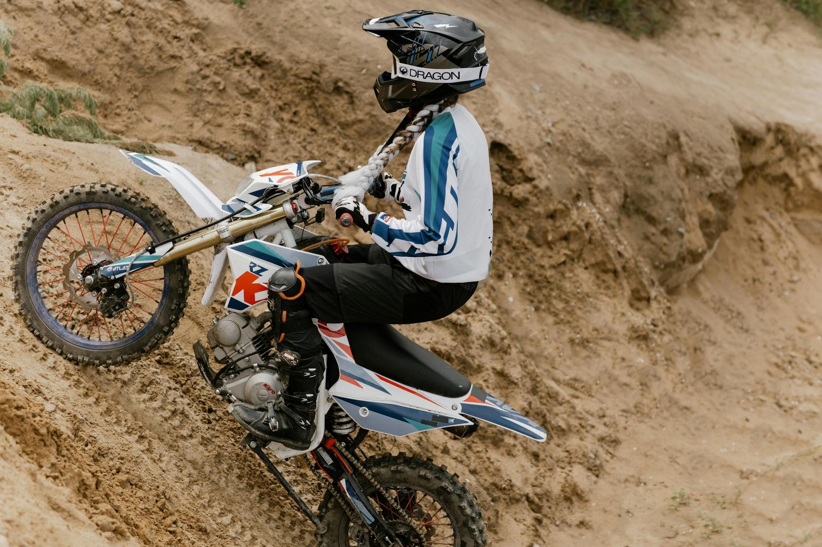 A motocross rider wearing protective gear ascends a sandy hill on a dirt bike during the day.