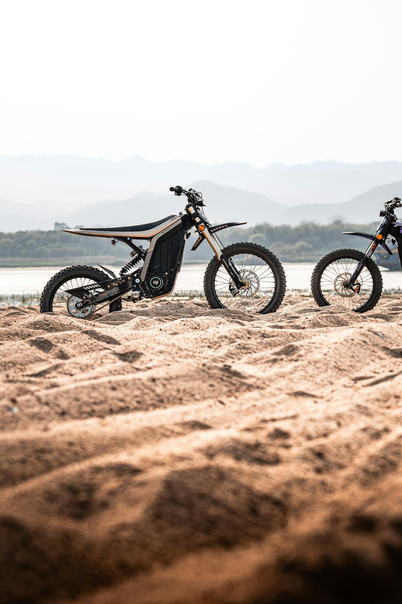 Electric off-road motorcycles on sandy terrain with distant mountains.