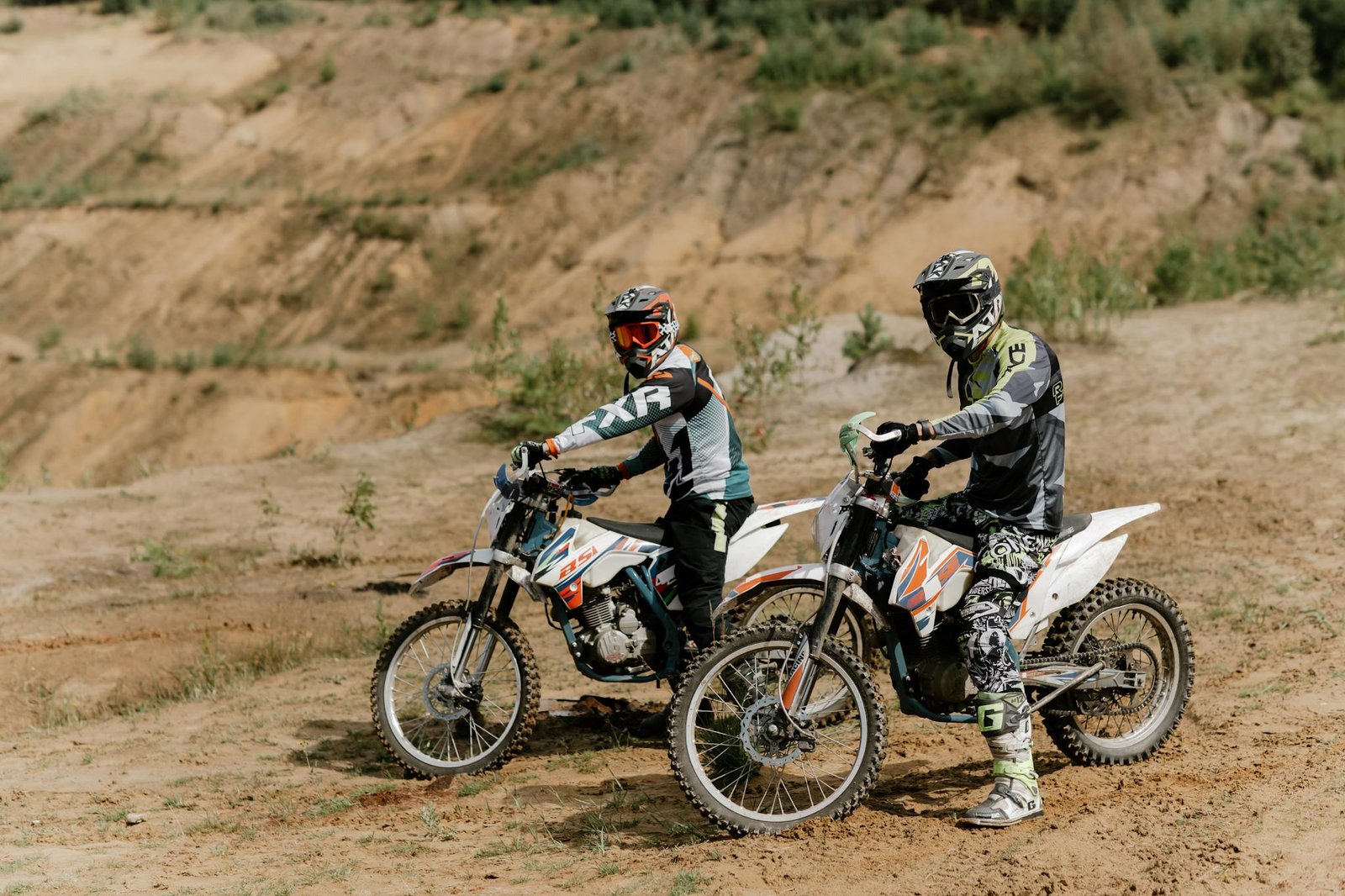 Two dirt bikers in protective gear pause on an off-road trail during a daytime ride.