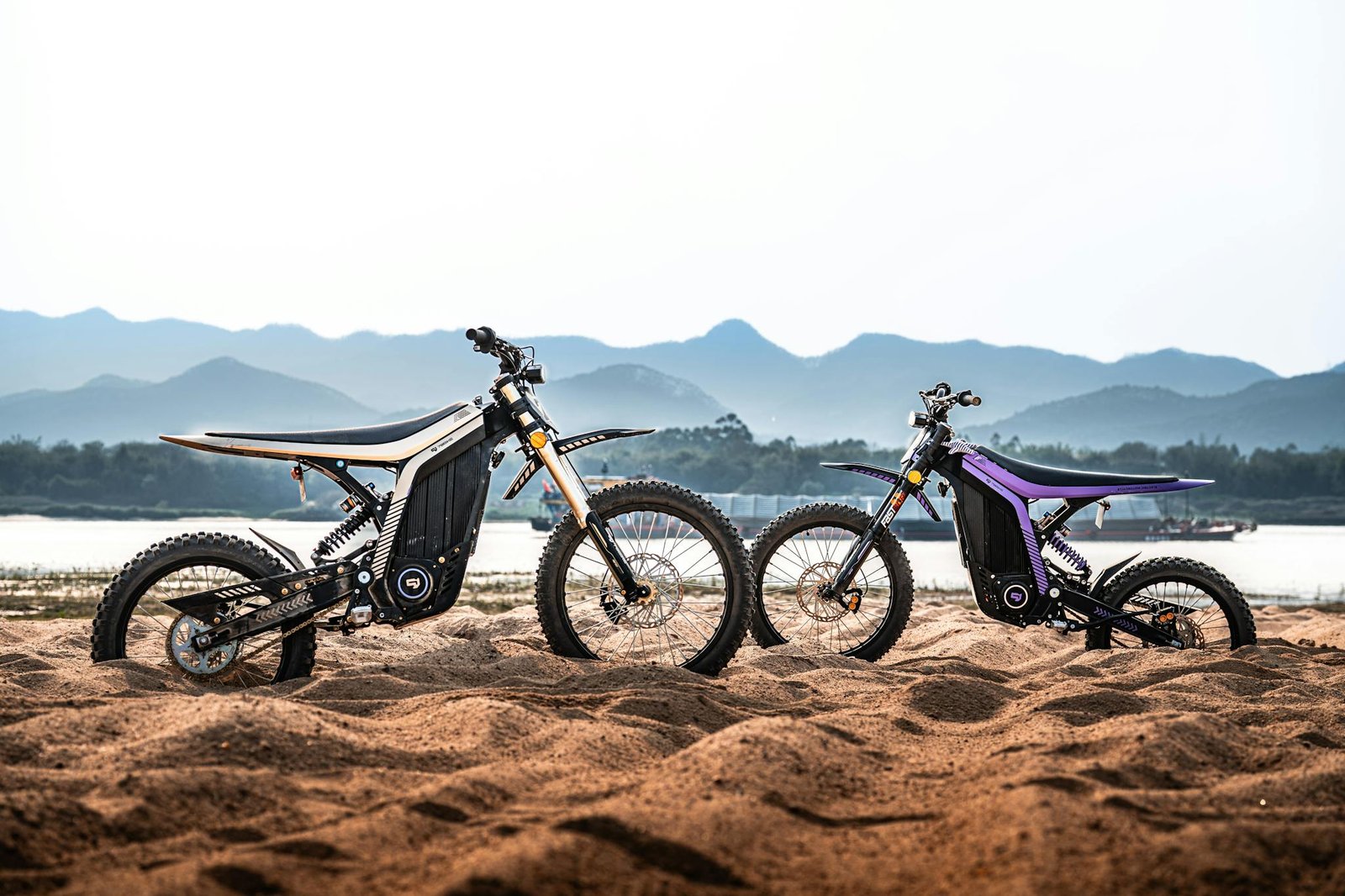 Two modern electric dirt bikes parked on a sandy beach with mountains in the background.